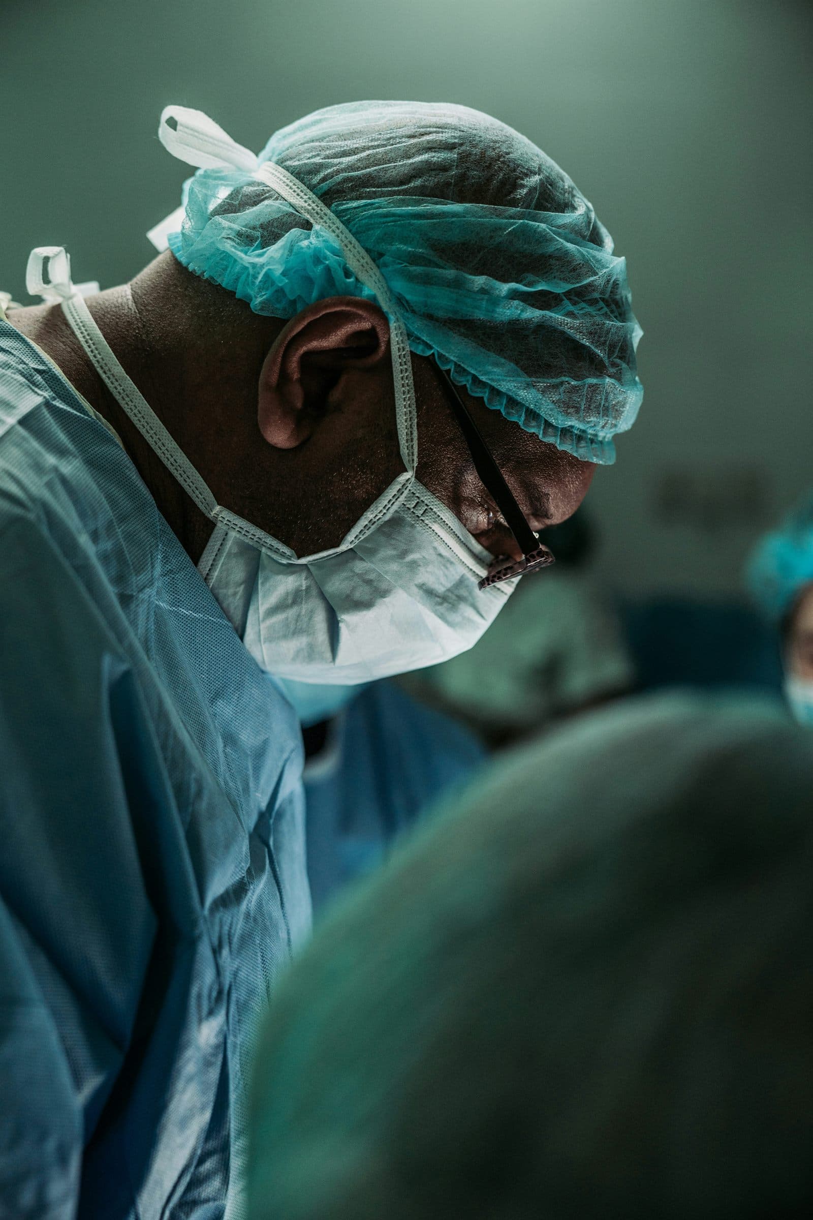 Surgeon in teal scrubs, cap, and mask leaning over a patient mid-operation