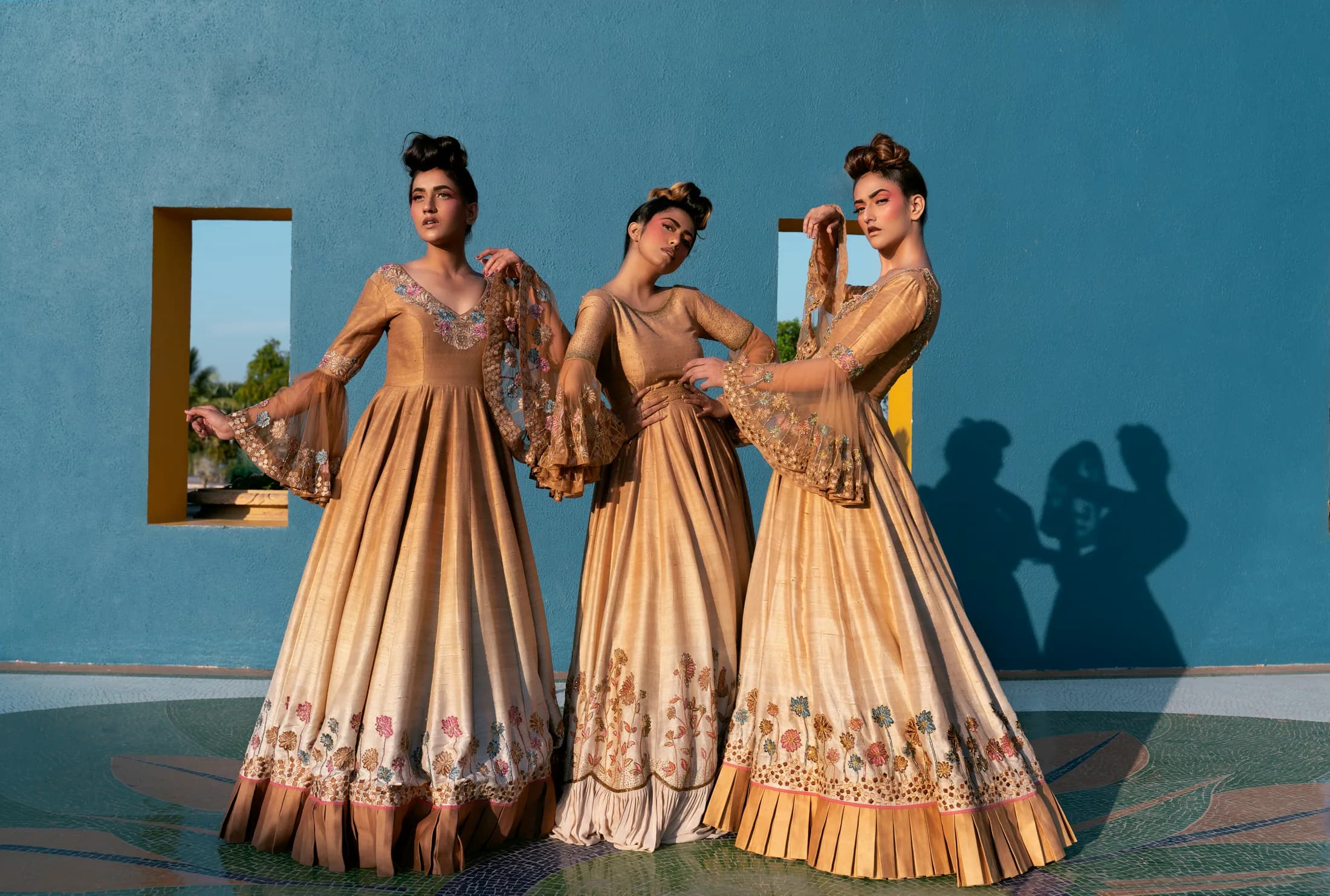 Three models in gold embroidered gowns against a blue wall