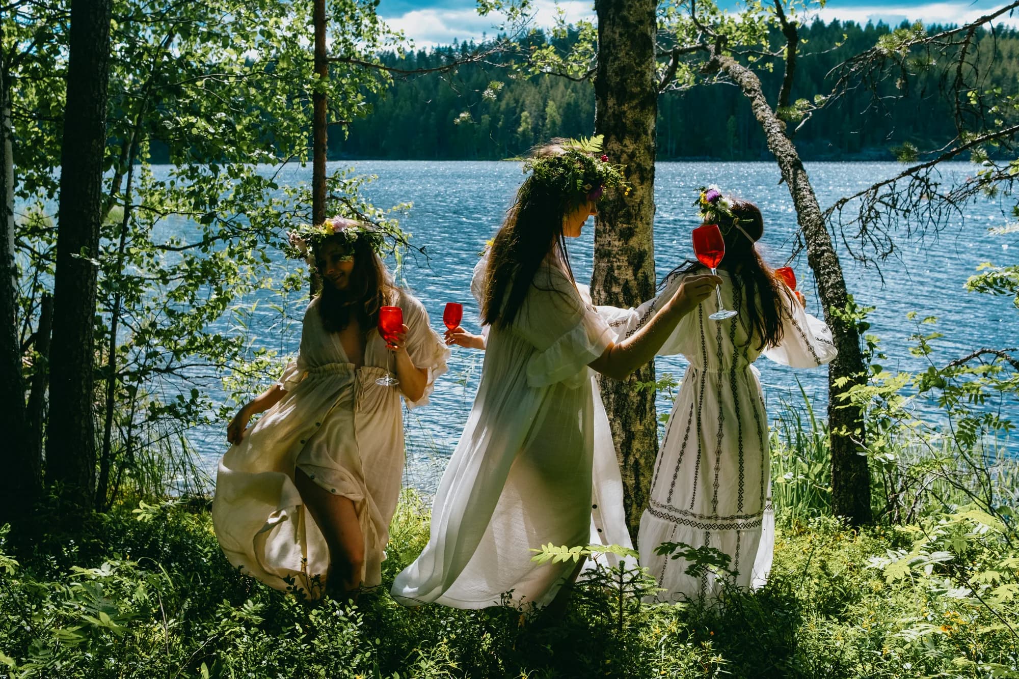 Three women in flowing white dresses and flower crowns by a forest lake