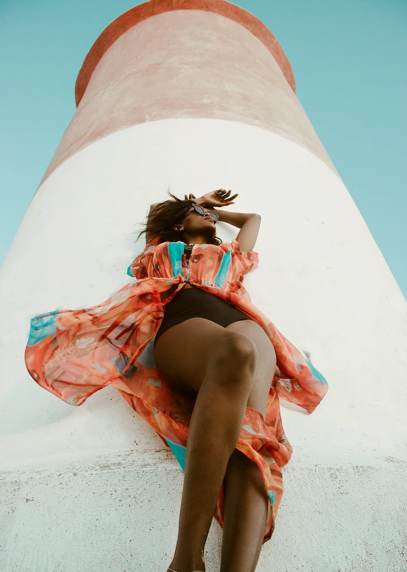 Model in a coral chiffon cover-up lying against a lighthouse under blue sky
